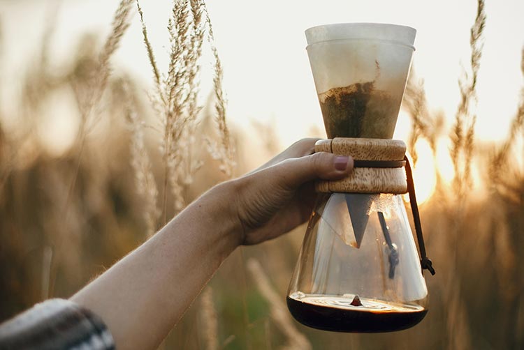 Hand mit heißem kaffee in glasflasche mit filter auf sonnigem, warmem licht in ländlichen kräutern. alternative kaffeebrauen im freien auf reisen. stimmungsvoller rustikaler ruhiger moment. platz kopieren