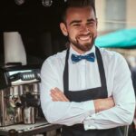 Gutaussehender barista, der in einem mobilen café im freien im handelszentrum der stadt arbeitet und eine schürze und ein weißes hemd mit fliege trägt, für die kamera posiert und lächelt