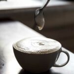 Tasse kaffee mit schaum, der von einer menschlichen hand gerührt wird.