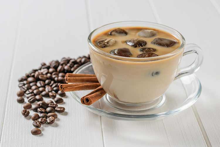Glasschale mit eiskaffee, zimtstangen und verstreuten kaffeebohnen auf weißem tisch. erfrischendes und belebendes getränk aus kaffeebohnen und milch.