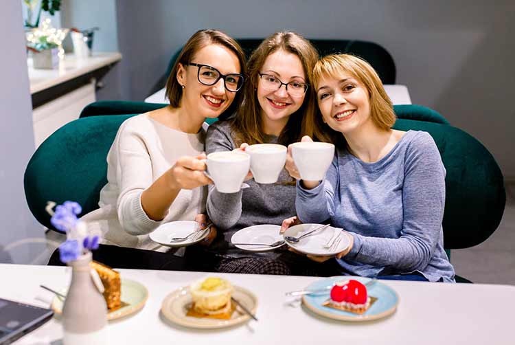 Three pretty Caucasian girl friends spend time together drinking coffee in the cafe, having fun and eating cakes and dessert.
