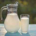 Frische milch wird in einen krug und ein glas gegossen, die auf einem türkisfarbenen holztisch vor dem hintergrund von grünem gras im sonnenlicht stehen. ziegenmilch ist sehr nützlich für kinder.