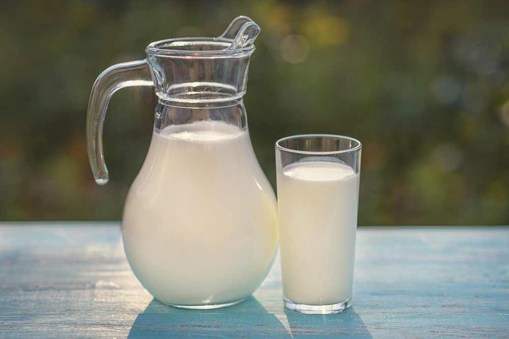 Frische milch wird in einen krug und ein glas gegossen, die auf einem türkisfarbenen holztisch vor dem hintergrund von grünem gras im sonnenlicht stehen. ziegenmilch ist sehr nützlich für kinder.