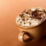 Tasse cappuccino mit schlagsahne und kaffeepulver auf beigem hintergrund.
