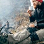 Horizontales bild im freien eines jungen mannes, der heißes getränk in den bergen in der nähe des lagerfeuers trinkt und sich nach dem trekking entspannt. reisender mit rotem hut, sitzt und hält nach dem wandern eine tasse tee. reisen