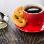 Kekse in form von brezeln, rote tasse mit kaffee auf untertasse, kondensmilch in schüssel, löffel auf holztisch