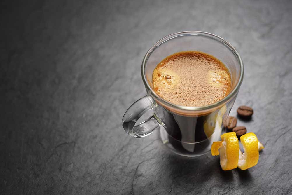 Transparente doppelwandige espresso-kaffeetasse aus glas mit zitronenschale auf schwarzem schieferhintergrund