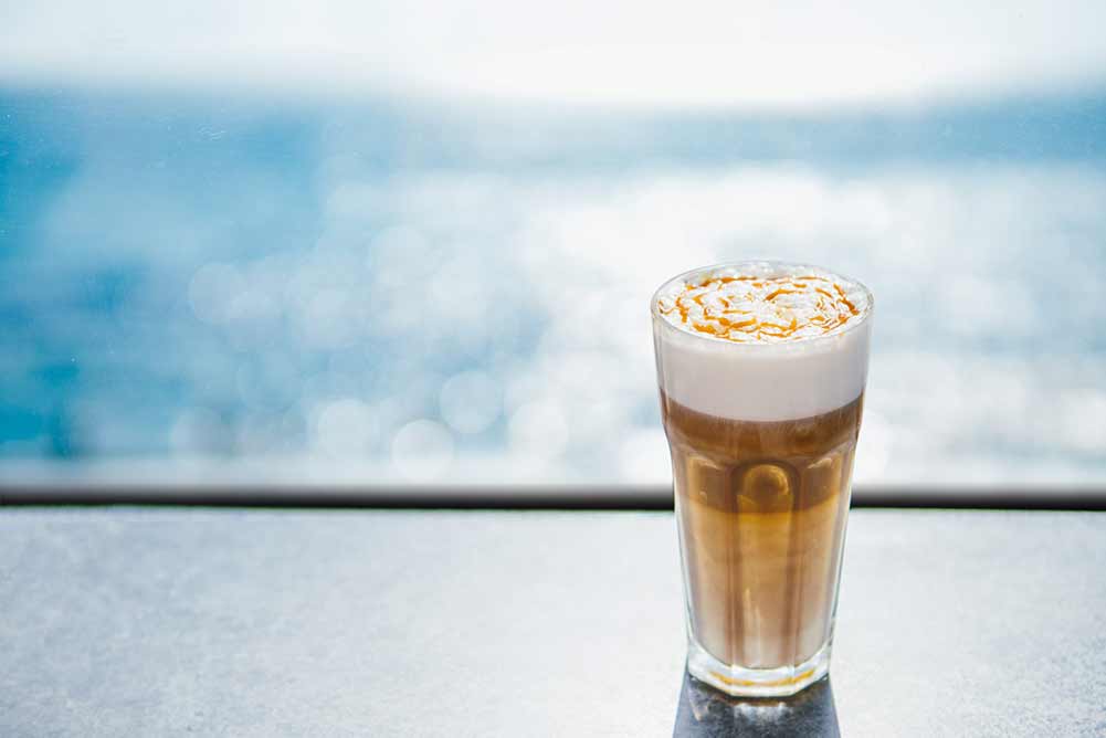 Heißer latte macchiato-kaffee mit karamell auf tischplatte mit blauem meer im hintergrund. natürliches hartes tageslicht. kopieren sie platz für text oder design