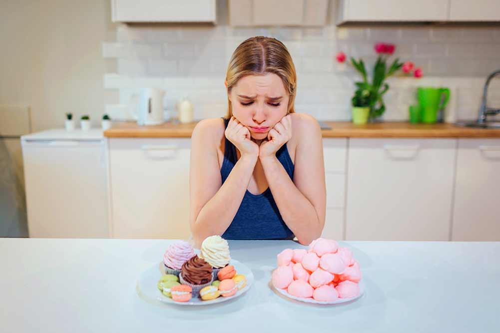 Diät kampf. junge traurige frau im blauen t-shirt wählt zwischen frischem obstgemüse oder süßigkeiten, während sie sie in der küche betrachtet. gesundes und ungesundes essen. diät