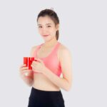 Schöne junge asiatische frau im sport nach dem training trinkwasser aus glas für durstig isoliert auf weiß