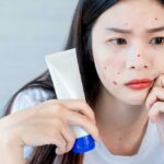 Asiatische teenager-frau, die spiegel betrachtet und akne-problem auf ihrem gesicht zusammendrückt, hautpflegekonzept.