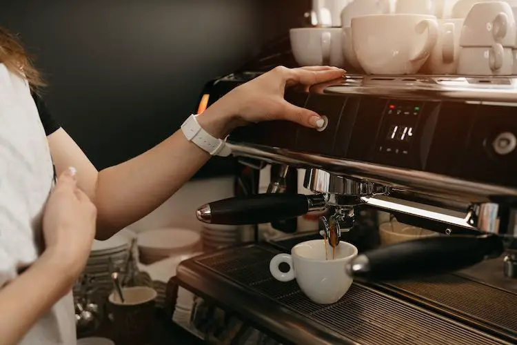 Ein weiblicher barista mit weißer schürze, der espresso in einer professionellen espressomaschine in einem café brüht. ein nahaufnahmefoto des eingießens eines kaffees in eine weiße tasse aus einer kaffeemaschine in einem café.
