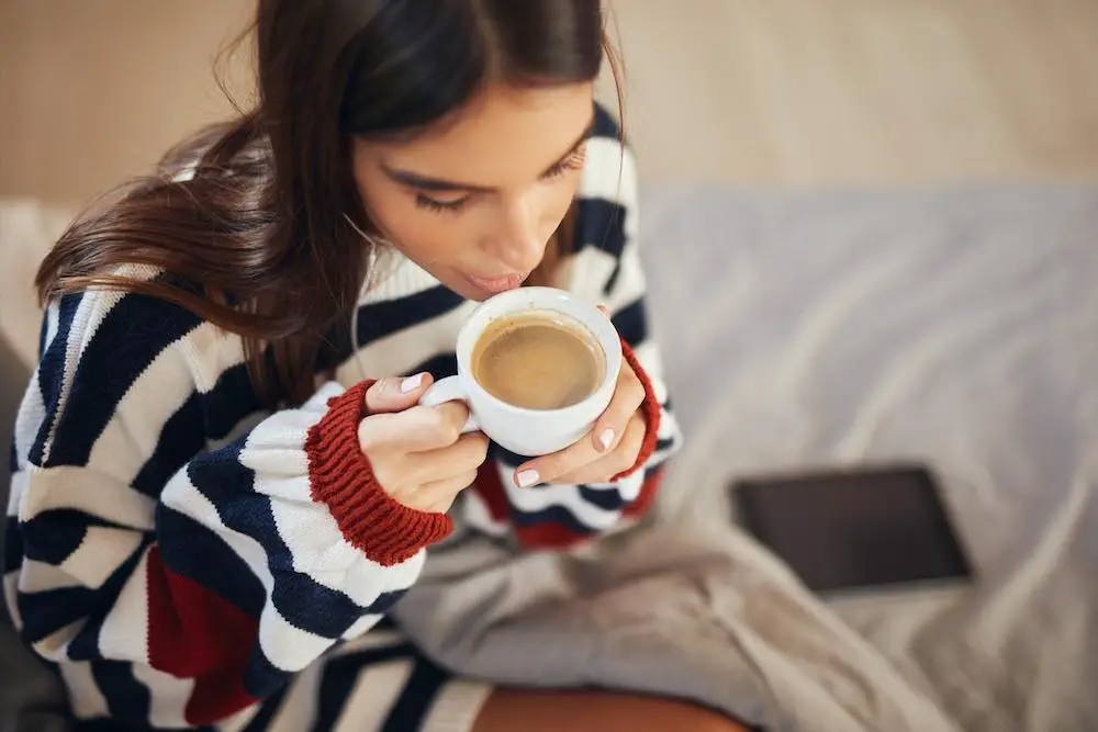 Draufsicht der schönen kaukasischen brünette im gestreiften pullover, die auf dem bett im schlafzimmer sitzt und ihren morgenkaffee trinkt.