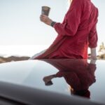 Frau sitzt mit kaffeetasse auf dem autodach und genießt den wunderschönen sonnenuntergang über den wolken