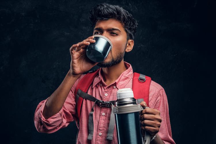 Junger stilvoller wanderer mit rucksack, der einen tee aus einer thermoskanne trinkt. studiofoto gegen eine dunkle strukturierte wand