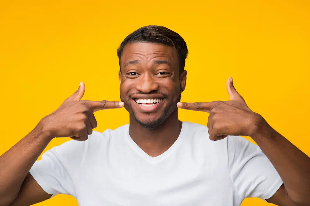 Fröhlicher afro-mann, der mit dem finger auf sein perfektes weißes lächeln zeigt, das auf gelbem hintergrund steht. studioaufnahme