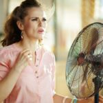Schwitzende junge hausfrau im modernen haus an sonnigen heißen sommertagen, die unter sommerhitze leiden, während sie vor dem ventilator steht.