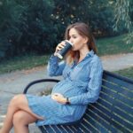 Glückliche gesunde mutterschaft. porträt einer schwangeren jungen kaukasischen frau, die draußen auf einer bank im park sitzt und kaffee aus einem pappbecher trinkt. ungesundes schlechtes essen während der schwangerschaft