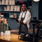 Freundlicher afrikanischer barista, der eine tasse kaffee hält, während er hinter einer theke in einem café steht und in eine kamera schaut