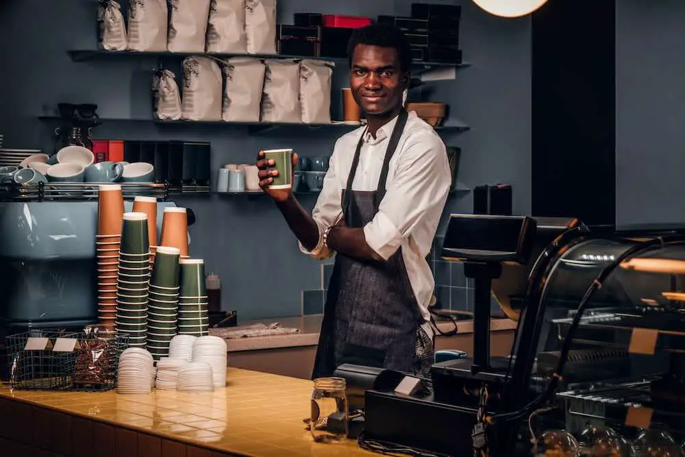 Freundlicher afrikanischer barista, der eine tasse kaffee hält, während er hinter einer theke in einem café steht und in eine kamera schaut