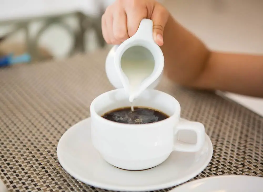 Heißer Kaffee mit einer Hand, die Milch auf Frühstückstisch gießt